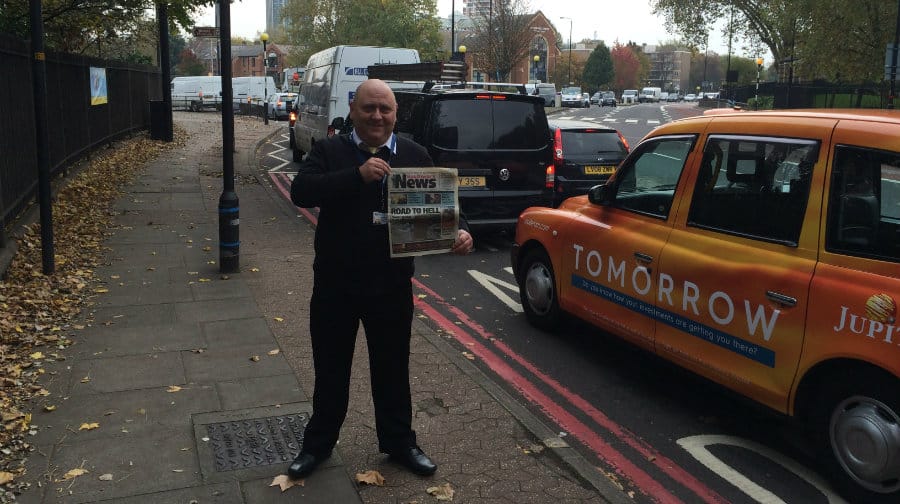 Dave Marks in front of Rotherhithe traffic, holding a copy of the Southwark News detailing his 2013 protest.