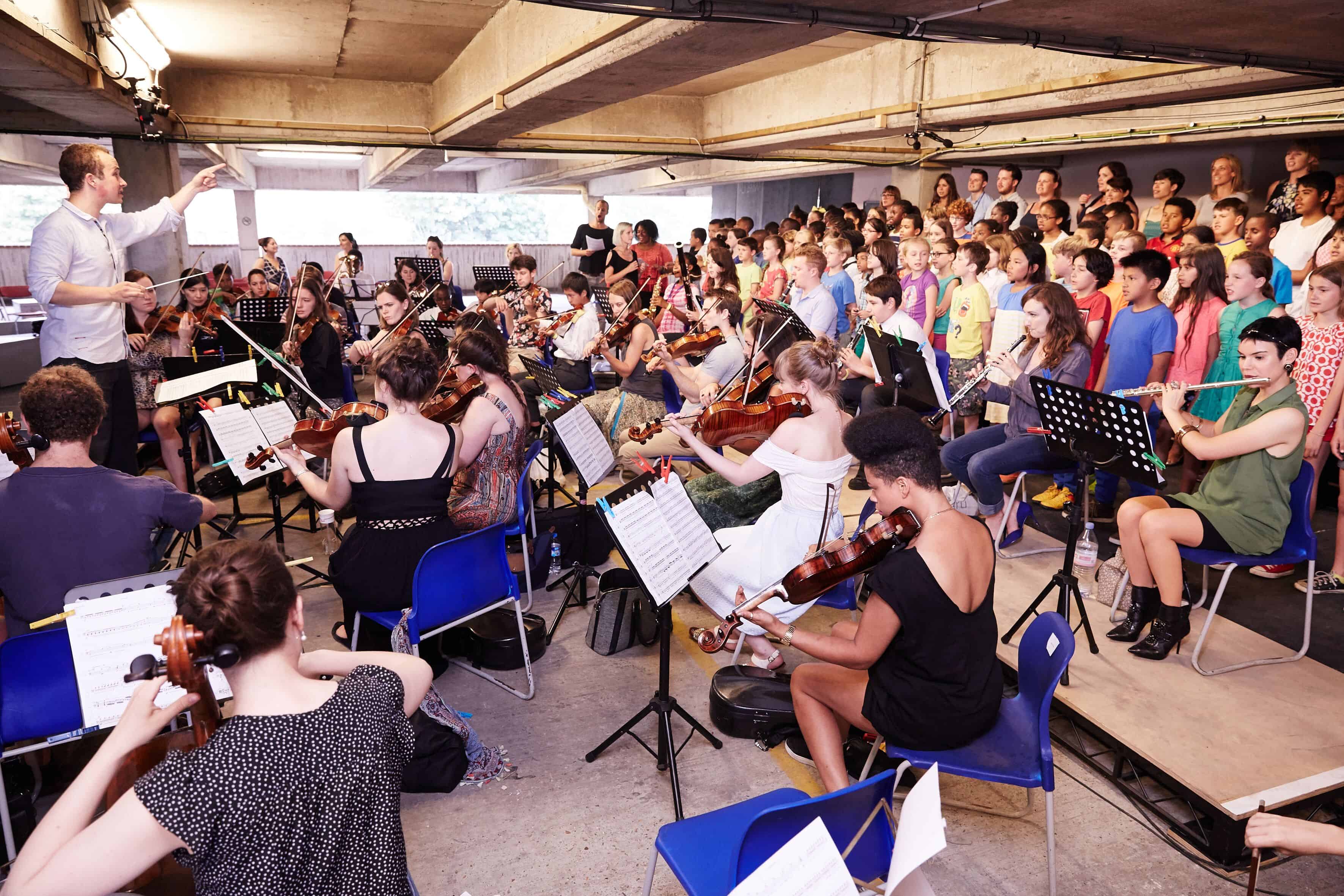 Composer and co-founder Christopher Stark leads an orchestra and youth choir at a previous car park performance.